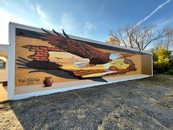 A large mural of an eagle souring through a hole in a brick wall. The mural appears 3 dimensional. Yje mural is 60 feet wide and 15 feet tall